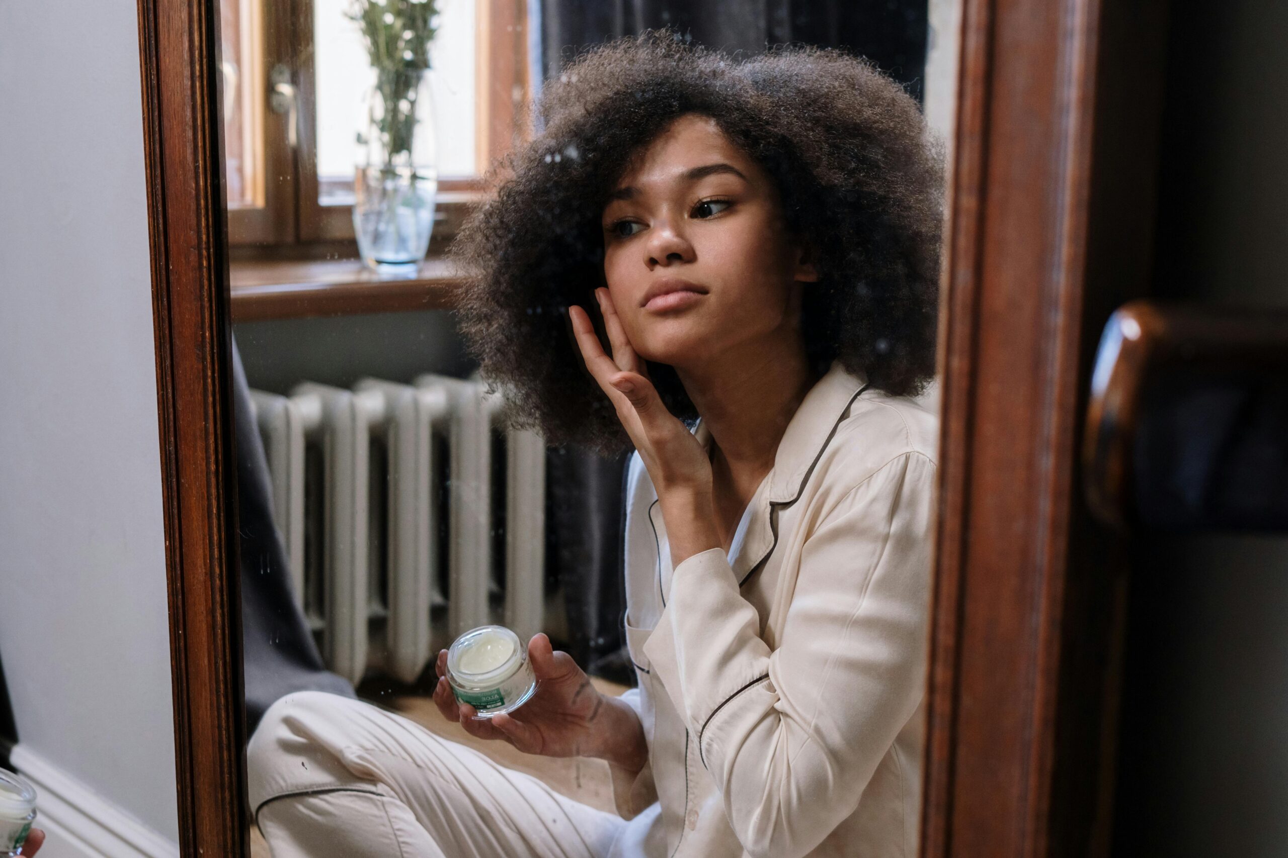 woman in cream pajamas applying skincare to her cheek, showing a glowing mirror skin routine.